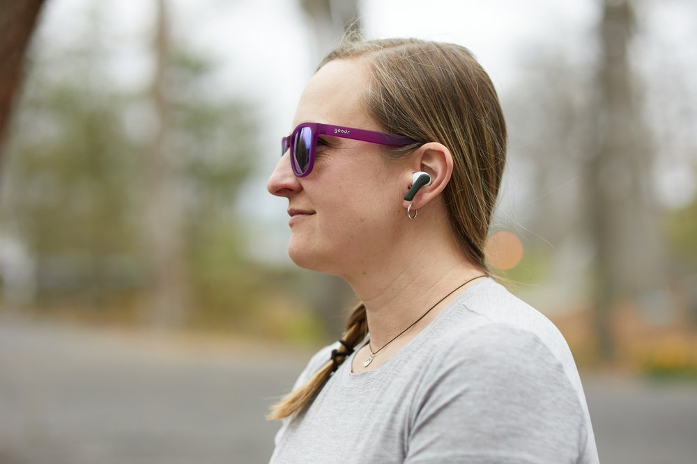 a girl with long hair, wearing purple sunglasses and a gray shirt, stands outdoors with a single earbud, looking left.