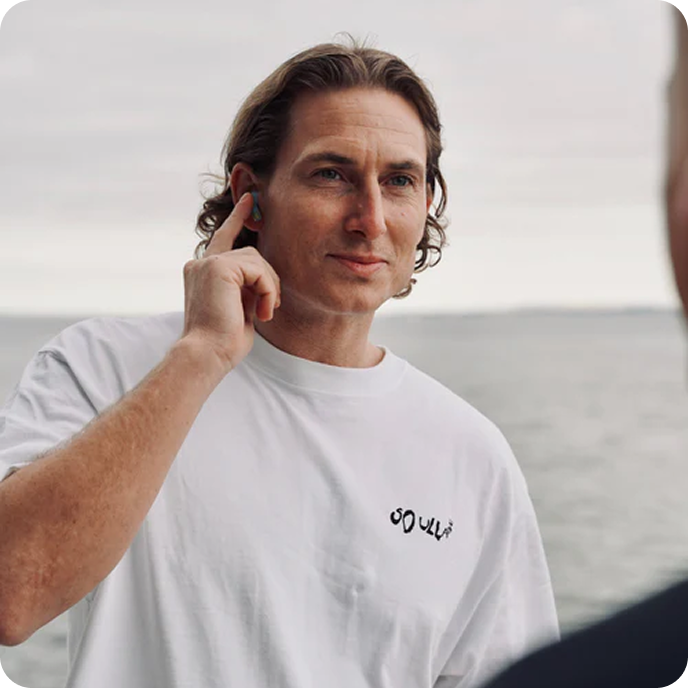 Man Using Touch Controls on Groovz Wireless Earbuds in Colorful Design – Speaking Outdoors Near Ocean in Casual White Tee