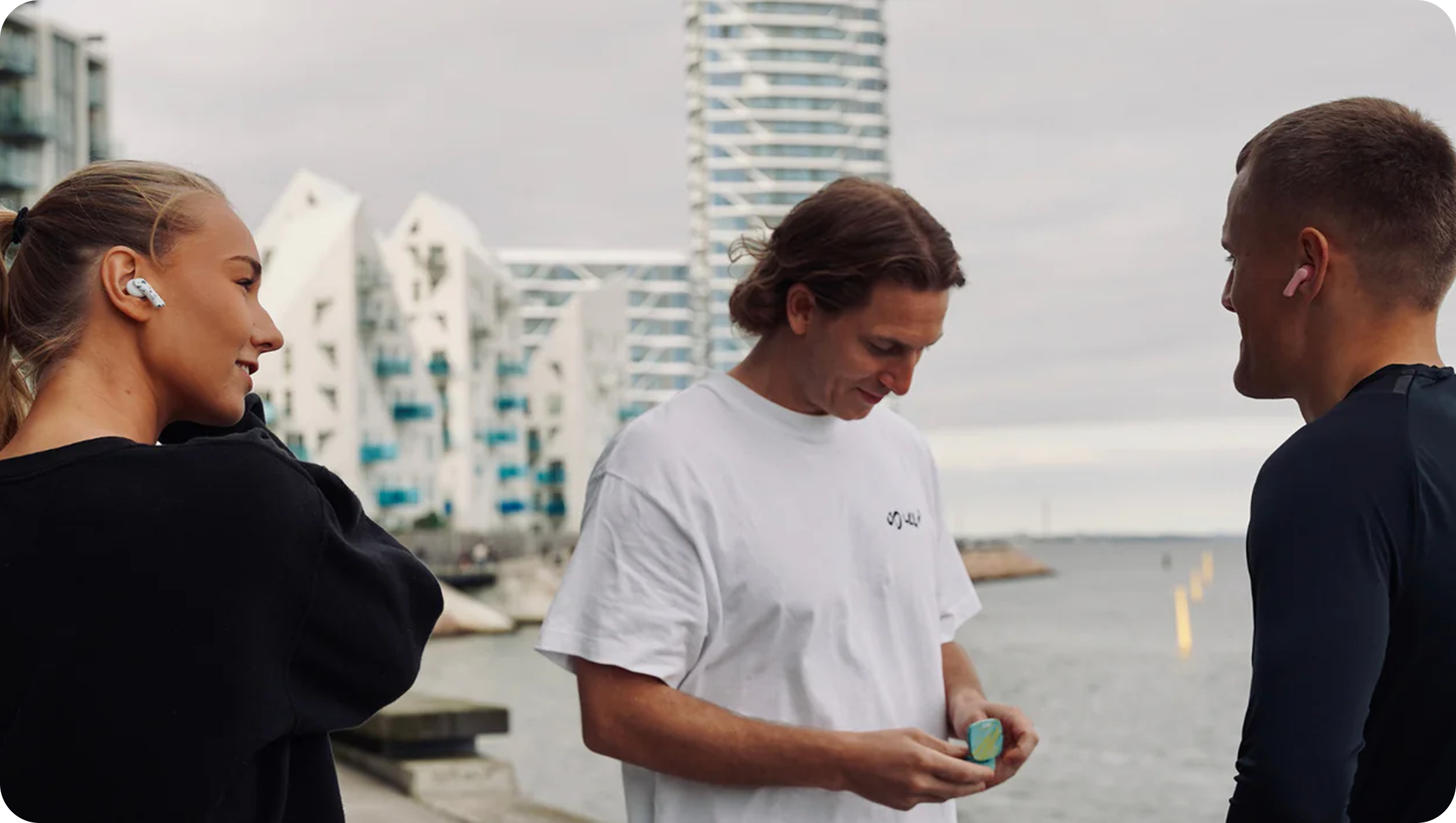 Three Friends Wearing Groovz True Wireless Earbuds in White, Pink, and Swirl Blue – Outdoor Lifestyle Scene on Waterfront with Modern Architecture
