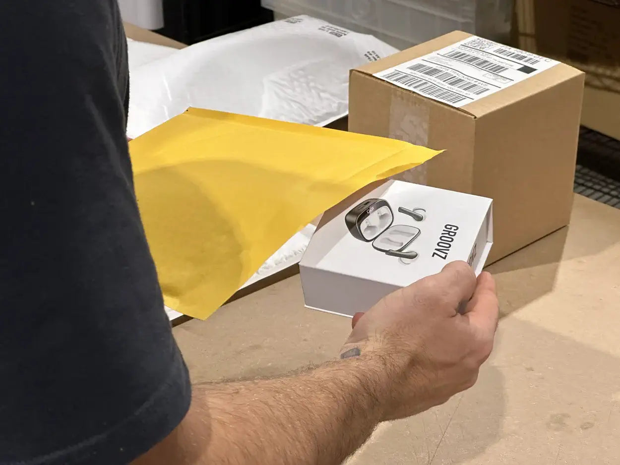 a person with a small tattoo is placing a white "Groovz" box into a yellow padded envelope, with a sealed brown package in the background.