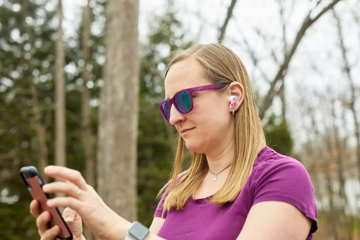 a lady in a purple top and sunglasses uses a smartphone outdoors, wearing floral earbuds. Trees are visible in the background.